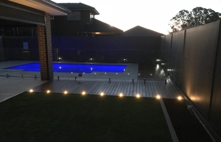 Backyard pool at dusk with composite decking, glass pool fence and ambient lighting in the lawn edge.